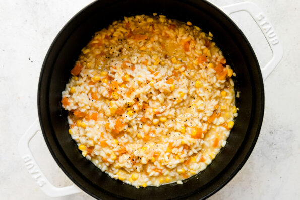 Finished sweet corn risotto fills a large white Staub Dutch oven that sits atop a creamy white textured surface. The risotto with corn has been garnished with fresh cracked pepper.
