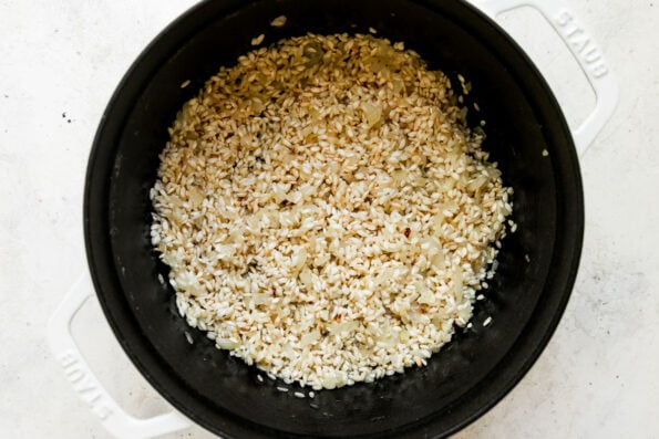 How to make corn risotto, step 3: Toast the arborio rice. Arborio rice, melted butter, sauteed onions and garlic fill a large white dutch oven. The rice toasts inside of the dutch oven and the dutch oven sits atop a creamy white textured surface.