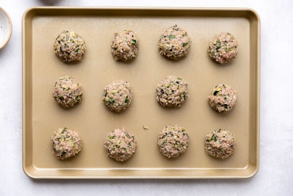 Twelve formed zucchini meatballs are arranged on a gold nonstick baking sheet sprayed with nonstick cooking spray. The baking sheet sits atop a creamy white textured surface surrounded by two small pinch bowls filled with kosher salt and ground black pepper.