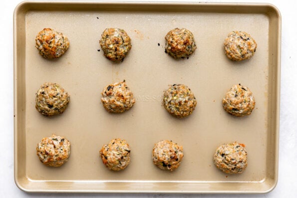 Twelve baked turkey zucchini meatballs are arranged on a gold nonstick baking sheet. The baking sheet sits atop a creamy white textured surface.