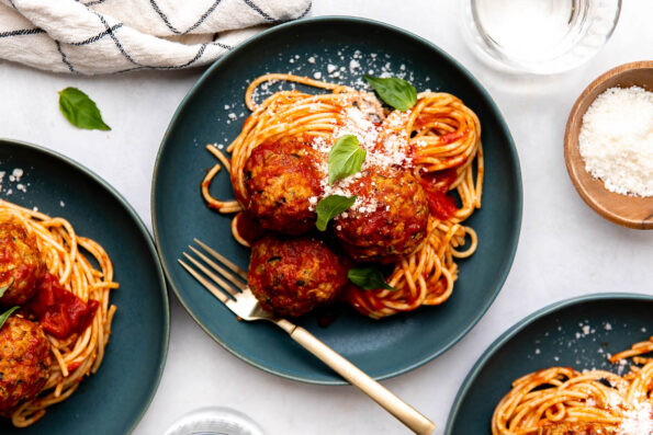 An overhead shot of three servings of zucchini meatballs with spaghetti and tomato sauce are served atop blue plates. The meatballs are garnished with fresh basil leaves and ground parmesan. Gold silverware rests atop the plate at center and each of the plates rest atop a creamy white textured surface. Two clear drinking glasses, a white linen napkin with a black window pane pattern, and a small wooden pinch bowl filled with grated parmesan surround the three servings of spaghetti and zucchini meatballs.