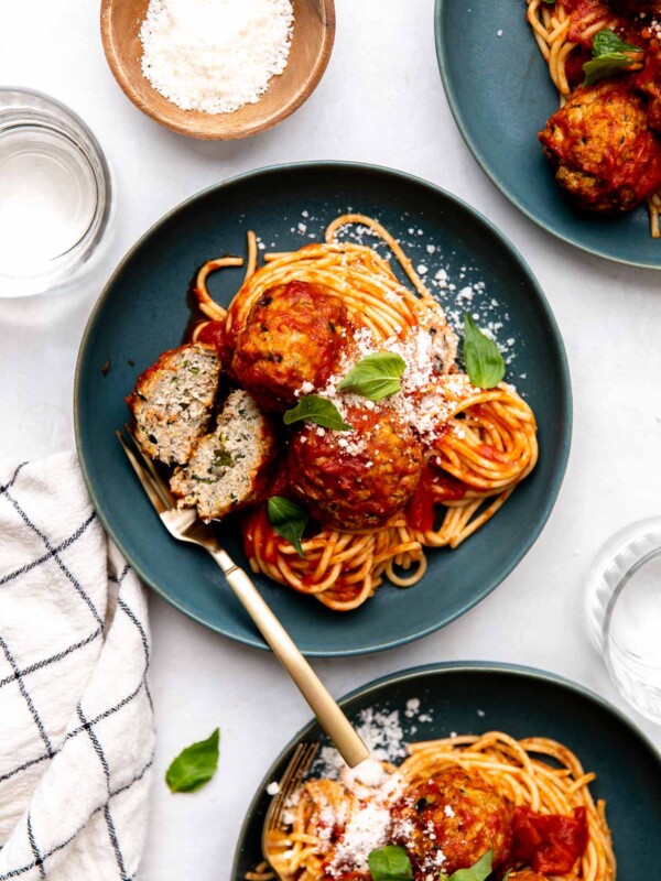 An overhead shot of three servings of zucchini meatballs with spaghetti and tomato sauce are served atop blue plates. The meatballs are garnished with fresh basil leaves and ground parmesan. Gold silverware rests atop the plate at center and each of the plates rest atop a creamy white textured surface. The center plate features one zucchini meatball that has been sliced in half to reveal the grated zucchini within the meatball. Two clear drinking glasses, a white linen napkin with a black window pane pattern, and a small wooden pinch bowl filled with grated parmesan surround the three servings of spaghetti and zucchini meatballs.