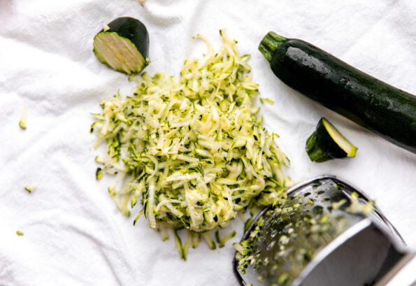 How to make a zucchini meatball recipe, step 2: Grate the zucchini. An overhead shot of a metal box grater used to grate zucchini rests atop a white linen towel. The ends of the grated zucchini as well as a full zucchini rest alongside.