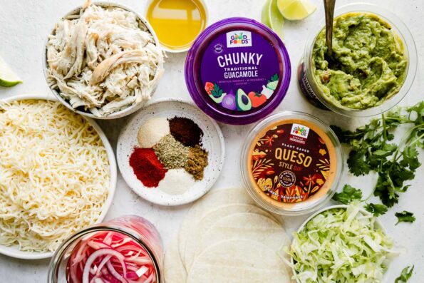 Baked crispy chicken taco ingredients arranged on a creamy white textured surface: shredded chicken breast, chili powder, ground cumin garlic powder, onion powder, dried oregano, smoked paprika, cayenne pepper, corn tortillas, olive oil, pepper jack cheese, Good Foods Chunky Guacamole, Good Foods Plant Based Queso Style Dip, pickled red onions, and shredded lettuce.