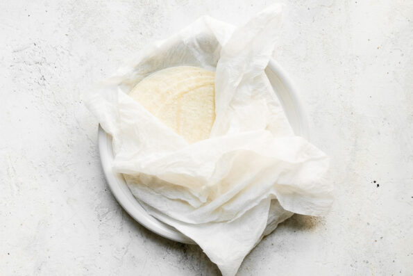 Corn tortillas wrapped in a damp paper towel rest atop a small white ceramic plate that sits atop a creamy white textured surface.
