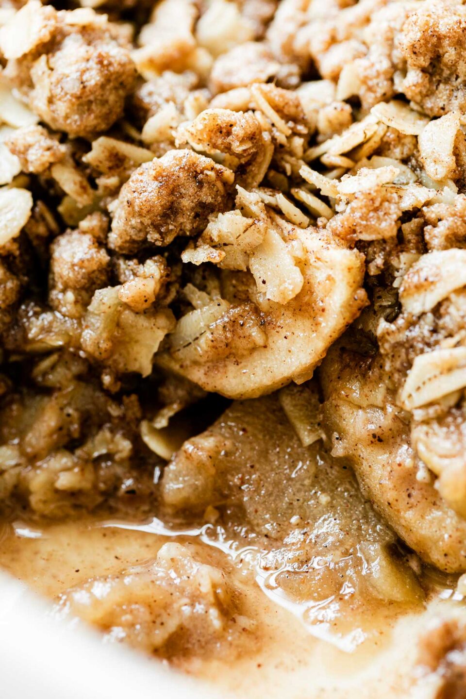 A close up and macro shot of spiced chai apple crisp inside of a white baking dish. A portion of the crisp has been scooped out revealing the layers of warm soft apples and browned chai spice crumble topping.