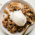 An overhead shot of spiced apple crisp with a scoop of vanilla ice cream inside of a white bowl with a dainty brown pattern. A gold fork rests inside of the bowl and the bowl sits atop a creamy white textured surface.