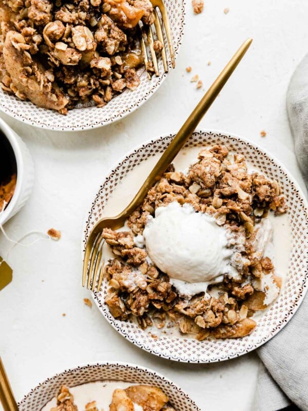An overhead shot of spiced apple crisp with a scoop of vanilla ice cream inside of a white bowl with a dainty brown pattern. A gold fork rests inside of the bowl and the bowl sits atop a creamy white textured surface surrounded by two other servings of apple crisp in a brown and white bowls, a gray linen napkin, and a small white ceramic cup of masala chai tea.