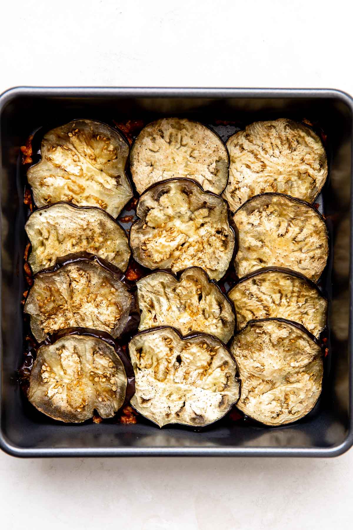 A layer of turkey bolognese topped with a layer of roasted eggplant rounds fills a 9x9 metal baking dish that sits atop a creamy white textured surface.