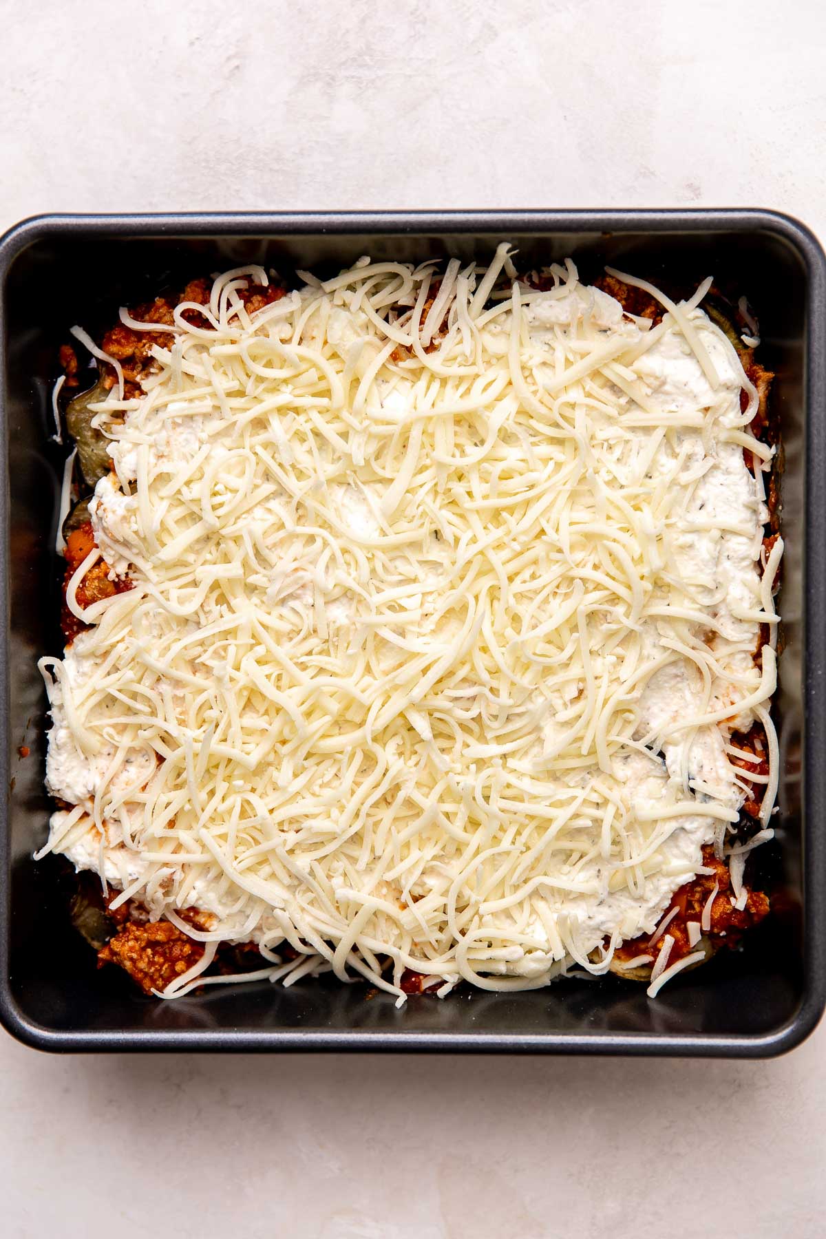 Another layer in the eggplant lasagna show the layer with roasted eggplant rounds topped with another layer of turkey bolognese, ricotta cheese mixture, and shredded mozzarella inside of a metal 9x9 baking dish. The baking dish sits atop a creamy white textured surface.