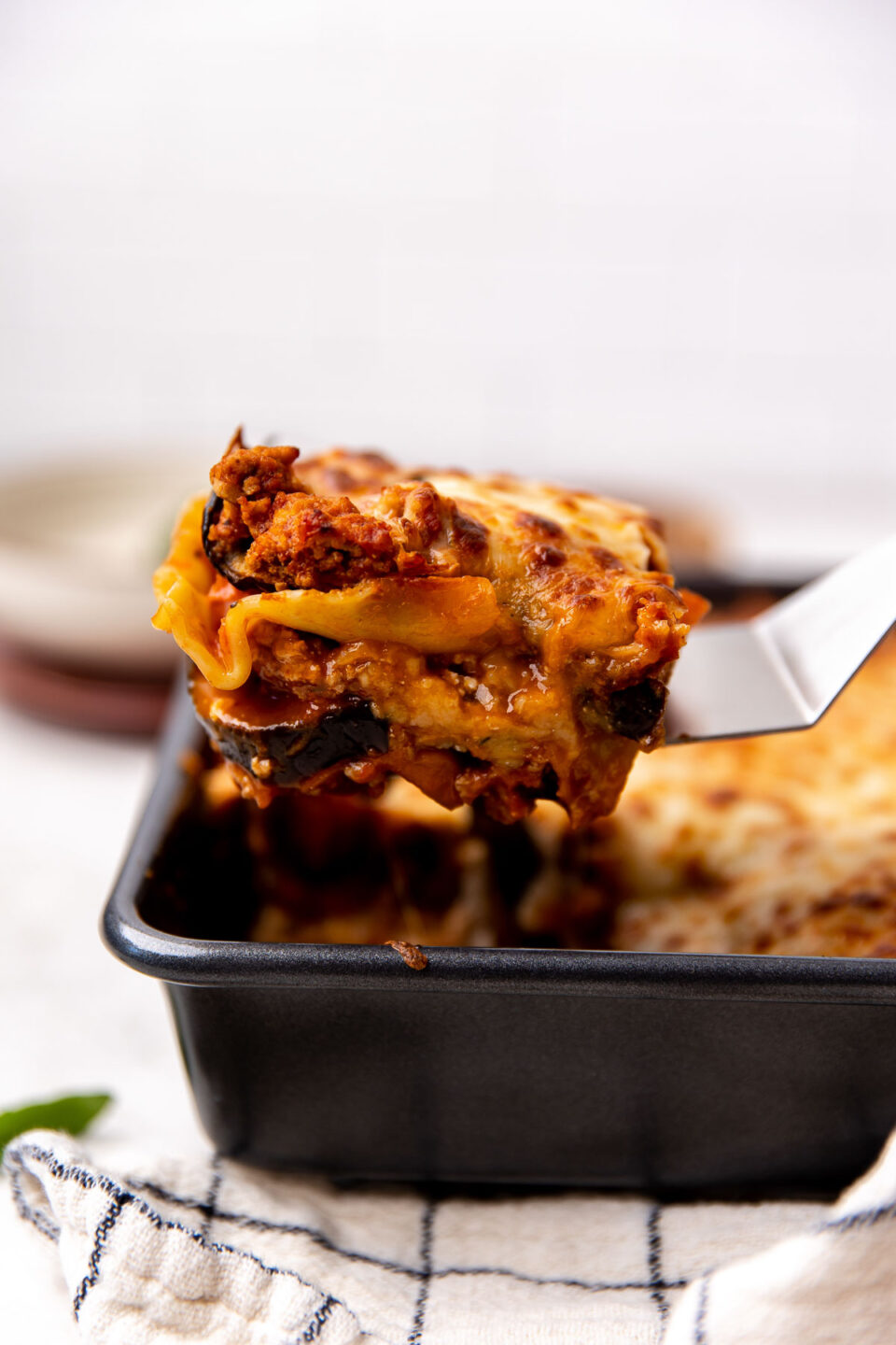 A single piece of eggplant lasagna is lifted out of a white baking dish with a metal spatula revealing the layers that make up the roasted eggplant lasagna and melty cheese.