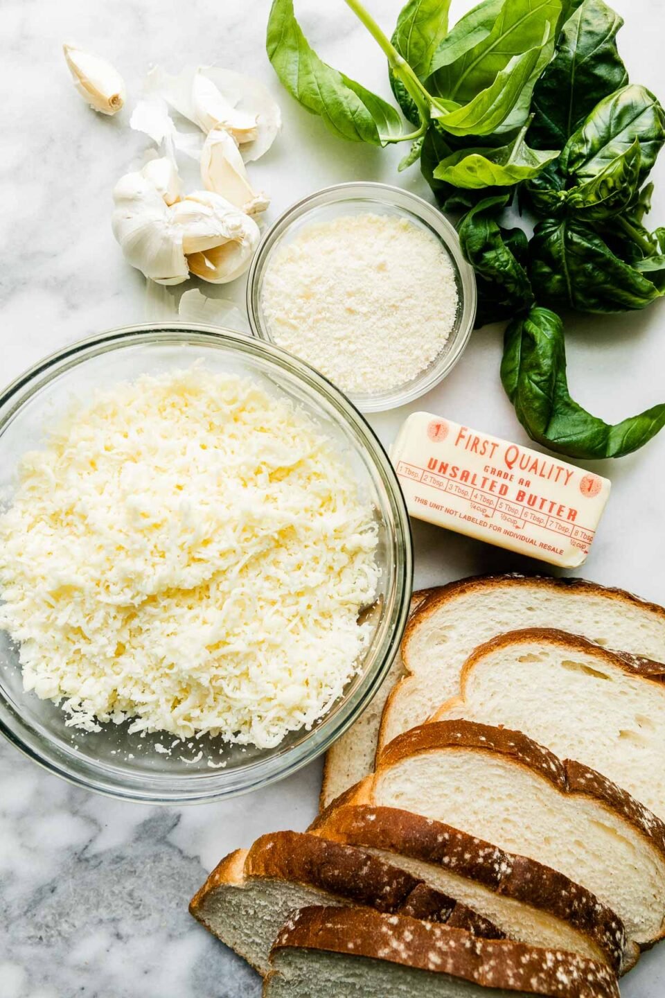 Mozzarella grilled cheese ingredients arranged on a white and gray marble surface: sliced bread choice, unsalted butter, garlic, finely grated parmesan, part-skim mozzarella, and fresh basil.