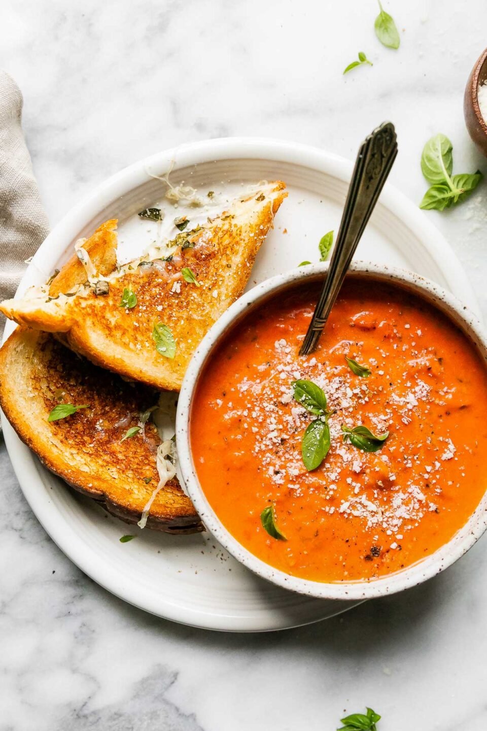 A grilled cheese with mozzarella, cut in half, rests atop a white ceramic plate alongside a small ceramic bowl filled with tomato soup. The sandwich and soup have been garnished with fresh grated parmesan and fresh basil. The plate sits atop a gray and white marble surface with a beige linen napkin and a small wooden pinch bowl filled with grated parmesan resting alongside.