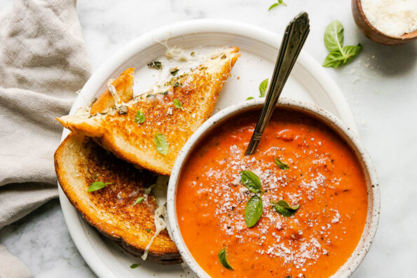 A mozzarella grilled cheese, cut in half, rests atop a white ceramic plate alongside a small ceramic bowl filled with tomato soup. The sandwich and soup have been garnished with fresh grated parmesan and fresh basil. The plate sits atop a gray and white marble surface with a beige linen napkin and a small wooden pinch bowl filled with grated parmesan resting alongside.