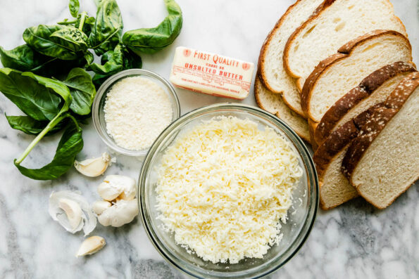 Mozzarella grilled cheese ingredients arranged on a white and gray marble surface: sliced bread choice, unsalted butter, garlic, finely grated parmesan, part-skim mozzarella, and fresh basil.