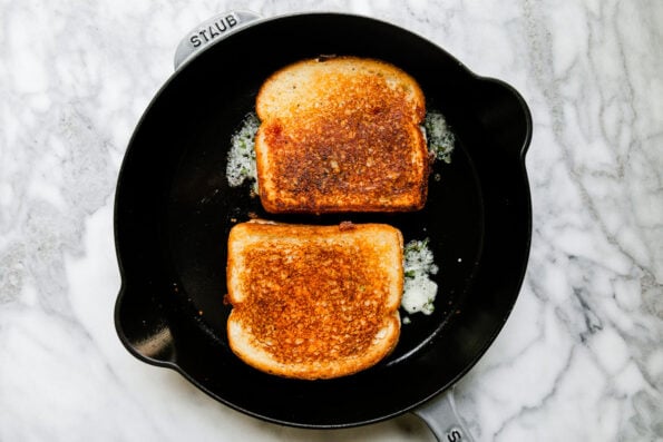 A large gray Staub cast iron skillet rests atop a gray and white marble surface. Two assembled mozzarella grilled cheese sandwiches cooked inside of the skillet.