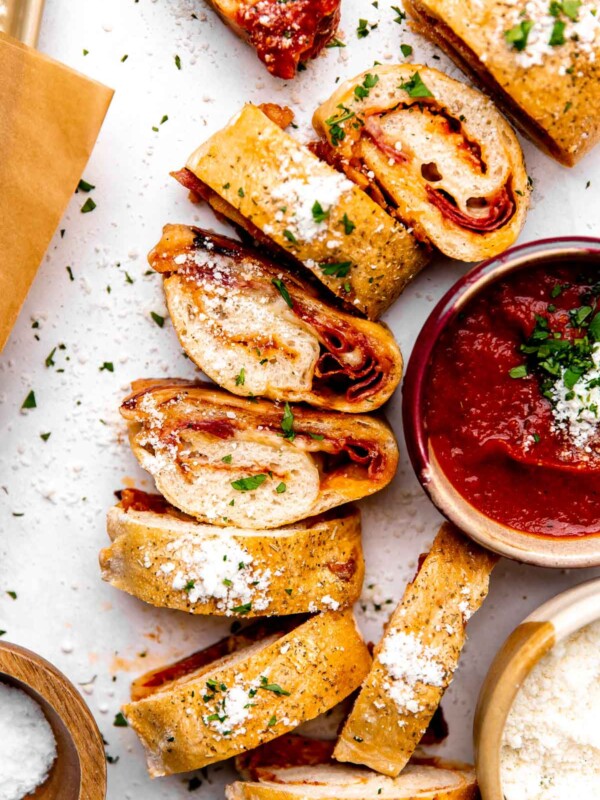 Sliced homemade stromboli pieces are arranged atop a creamy white textured surface. The slices have been garnished with fresh chopped herbs and grated parmesan cheese. A small ceramic bowl filled with DeLallo Pizza sauce, a small ceramic bowl filled with grated parmesan cheese, a small wooden pinch bowl filled with kosher salt, and a parchment lined baking sheet surround the sliced stromboli at center.