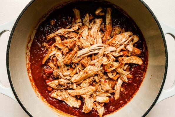Shredded chicken thighs are added back into a simmered chicken ragu sauce that fills a large cream colored Dutch oven that sits atop a creamy white textured surface.