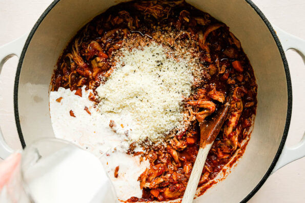 A woman's hand pours heavy creamy from above into a a large cream colored Dutch oven filled with chicken ragu sauce with grated parmesan added. The Dutch oven sits top a creamy white textured surface and a wooden spoon rests inside of the pot for stirring.