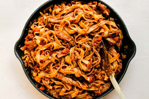 Chicken Ragu pasta fills a large white Le Creuset cast iron skillet that sits atop a creamy white textured surface. A wooden spoon rests inside of the skillet for stirring.
