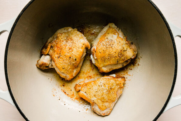 Brown chicken thighs rest inside of a large cream colored pan that sits atop a creamy white textured surface.