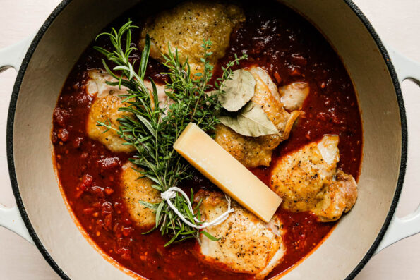 The beginnings of a chicken ragu sauce fill a large cream colored pan that sits atop a creamy white textured surface. At the top of the sauce there are browned chicken thighs nestled into tomatoes, a bunch of fresh herbs, bay leaves, and a parmesan rind.