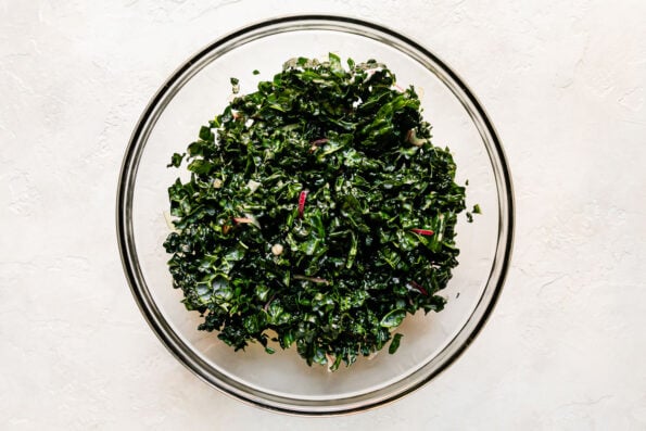 Kale massaged in lemon juice fills a large clear glass mixing bowl that sits atop a creamy white textured surface.