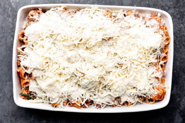 A final assembled but not yet baked, baked rigatoni fills a large white ceramic baking dish that sits atop a dark gray textured surface. The final layer of the rigatoni bake is a sprinkling of grated parmesan cheese.