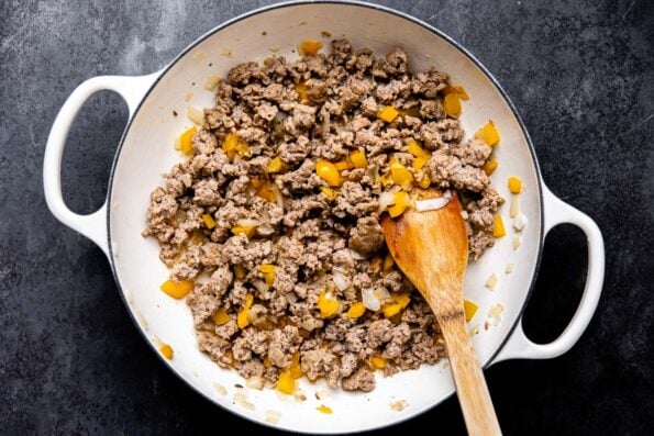 Browned spicy Italian sausage, bell peppers and onions fill a large white double handled pan that sits atop a dark gray textured surface. A wooden spoon rests inside of the mixture for stirring.