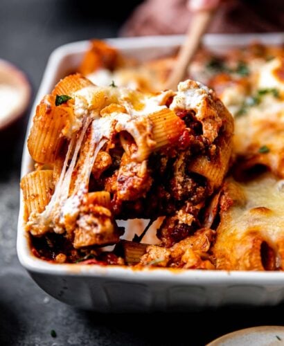 A side angle shot of a scoop of baked rigatoni with sausage being taken out of a large white ceramic baking dish with a wooden serving spoon. The baking dish sits atop a dark gray textured surface surrounded by small pinch bowls.