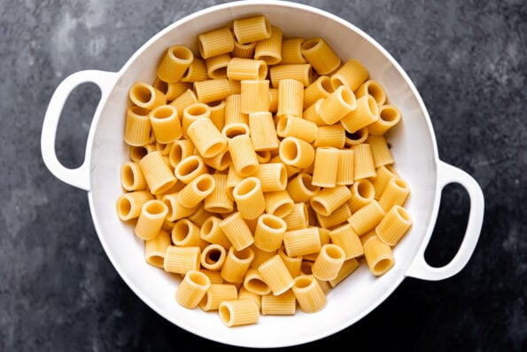 DeLallo mezzi rigatoni pasta cooked just a few minutes under al dente is strained inside of a white metal colander that sits atop a dark gray textured surface.