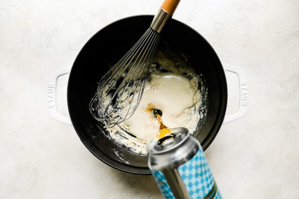 A large white Staub cocotte is filled with a roux while a can of beer is slowly poured into it. A metal whisk rests inside of the pan for stirring. The pot sits atop a creamy white textured surface.