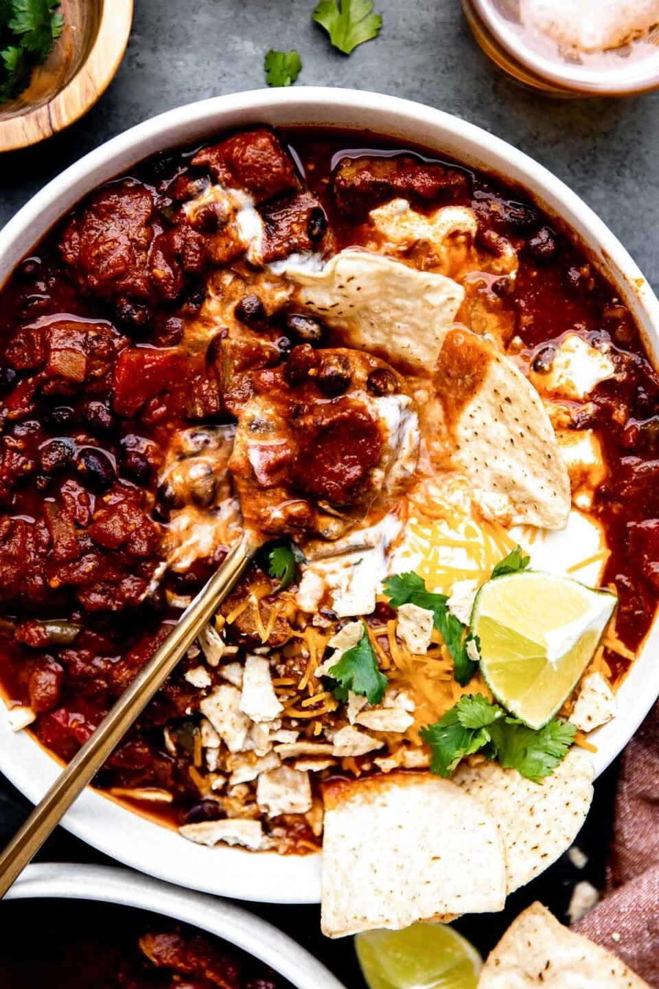 Beef chili is served inside of a white bowl that sits atop a dark textured surface. The chili with stew meat has been topped with a dollop of sour cream, whole and crushed tortilla chips, fresh cilantro, shredded cheese and a lime wedge. A gold spoon rests inside of the bowl with a spoonful of chili scooped on top. The bowl is surrounded by a burnt orange colored linen napkin, loose fresh cilantro, a cut lime half, loose tortilla chips, an amber colored drinking glass, and a second white bowl filled with beef chili.