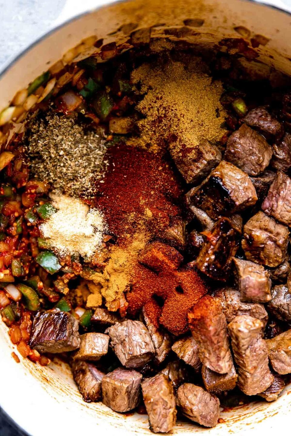 How to make a braised beef chili, step 3: Build the beef chili & braise. Browned beef, vegetables, and chili spices are added into a large pot that sits atop a dark textured surface to begin to build chili with stew meat.
