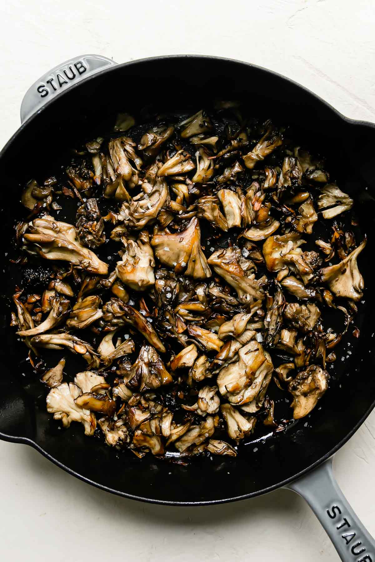 Pan-roasted mushrooms fill a light gray Staub cast iron skillet. The skillet sits atop a creamy white textured surface.