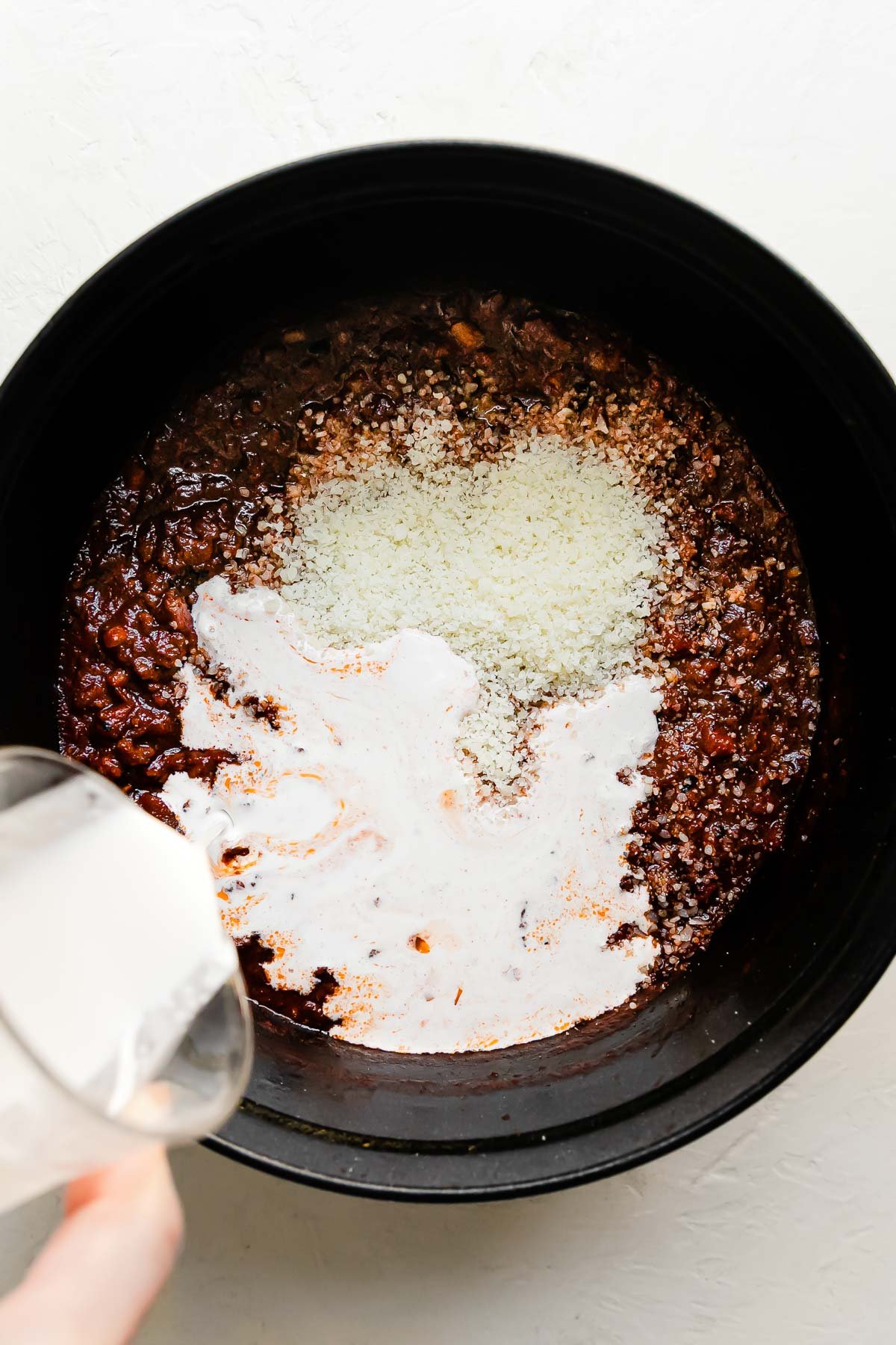 Grated parmesan and bolognese sauce fill a large white Staub cocotte that sits atop a creamy white textured surface. A woman's hand holds a glass of heavy cream above the cocotte as it is slowly poured into the bolognese sauce.