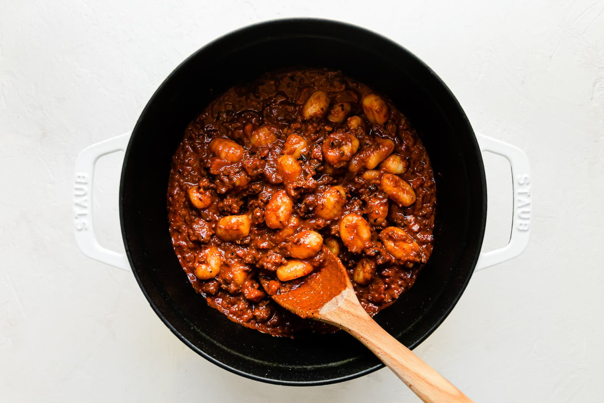 Gnocchi and bolognese sauce are tossed together inside of a large white Staub cocotte that sits atop a creamy white textured surface. A wooden spoon rests inside of the Dutch oven for stirring.