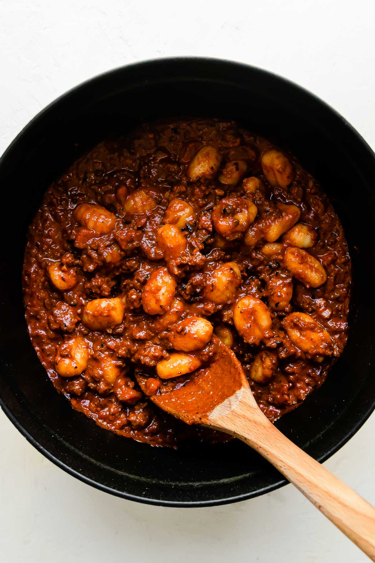 Gnocchi and bolognese sauce are tossed together inside of a large white Staub cocotte that sits atop a creamy white textured surface. A wooden spoon rests inside of the Dutch oven for stirring.