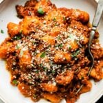 A plateful of gnocchi bolognese sits atop a creamy white textured surface. A dark gray linen napkin and two plates rest alongside the plate at center. A silver fork rests atop the plate of gnocchi.