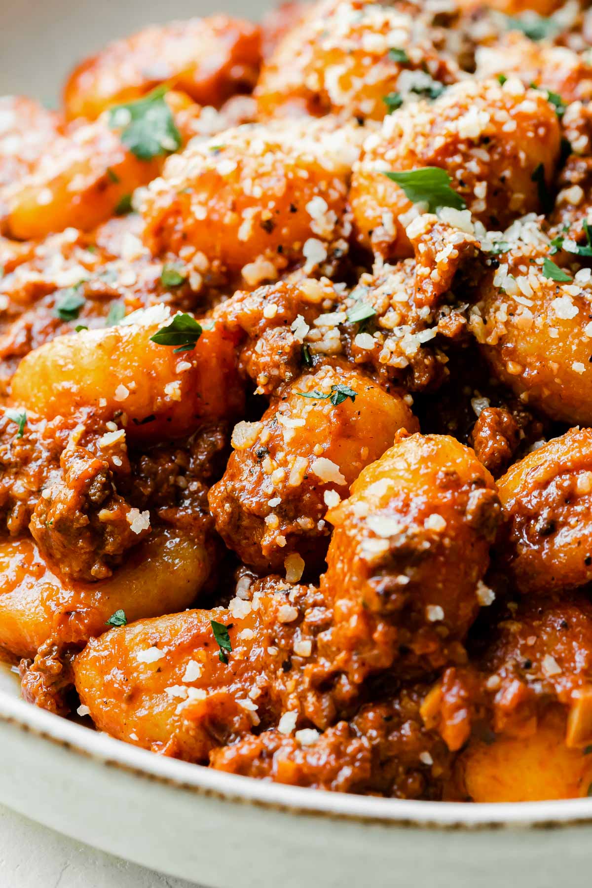 A close up and side angle shot of gnocchi bolognese atop a white ceramic plate that sits atop a creamy white textured surface. The bolognese gnocchi is garnished with grated parmesan and fresh chopped herbs.