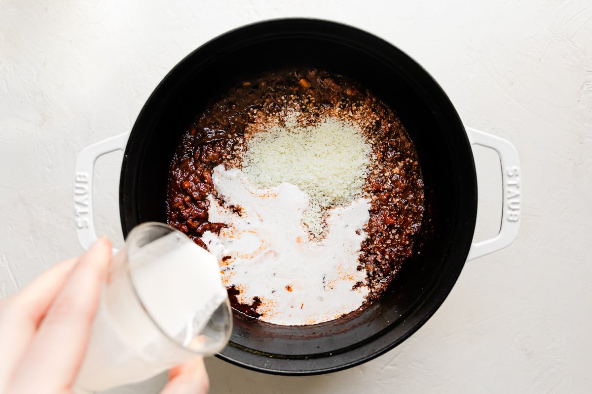 Grated parmesan and bolognese sauce fill a large white Staub cocotte that sits atop a creamy white textured surface. A woman's hand holds a glass of heavy cream above the cocotte as it is slowly poured into the bolognese sauce.