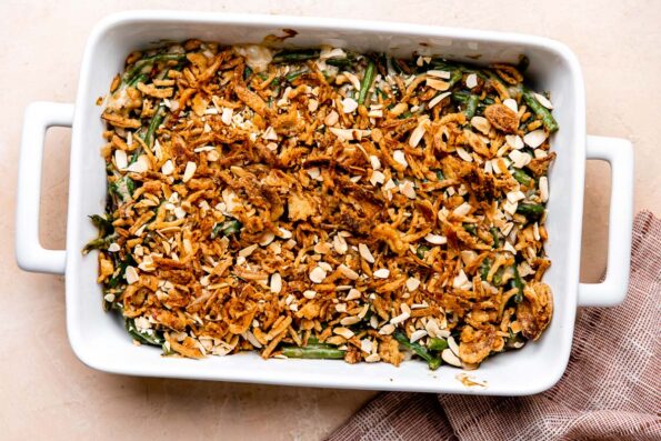 Finished green bean casserole made from scratch fills a large white ceramic baking dish that sits atop a light peach colored textured surface. A muted red linen napkin rests alongside the baking dish.