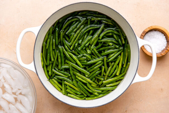 Par-cooked green beans in water, for green bean casserole with fresh green beans, fill a large white double handled pot. The pot sits atop a light peach colored textured surface surrounded by a large glass mixing bowl filled with ice water and a small wooden pinch bowl filled with kosher salt.
