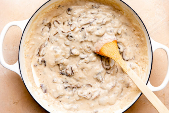 Mushroom sauce for make ahead green bean casserole fills a large white double handled braising pan that sits atop a light peach colored textured surface. A wooden spoon rests inside of the pan for mixing.