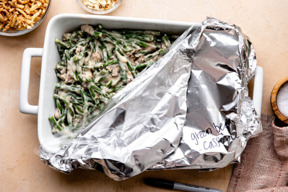 Partially assembled make ahead green bean casserole fills a large white ceramic baking dish that sits atop a light peach colored textured surface. The baking dish is partially covered with plastic wrap and aluminum foil and the words green bean casserole have been written on the outside of the foil in black permanent marker. The baking dish is surrounded by a black Sharpie marker, a bowl filled with French fried onions, a small wooden pinch bowl filled with kosher salt, another bowl filled with slivered almonds, and a red muted linen napkin.