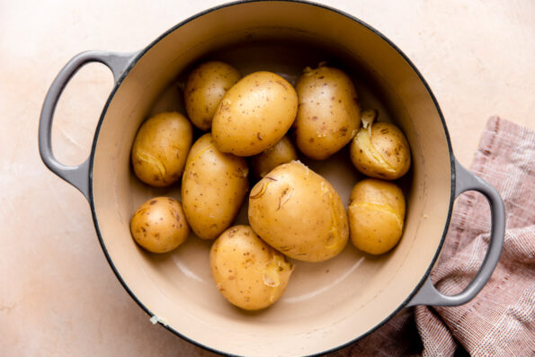 Boiled Yukon Gold potatoes that have been drained sit inside a gray double handled ceramic pot. The pot sits atop a light peach colored textured surface with a red muted linen napkin resting alongside.