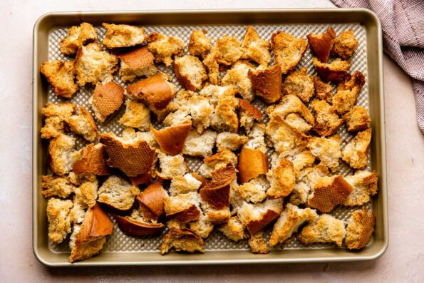 Roughly torn toasted bread cubes are arranged atop a metal baking sheet pan. The pan sits atop a light peach colored textured surface with a red muted linen napkin resting alongside.