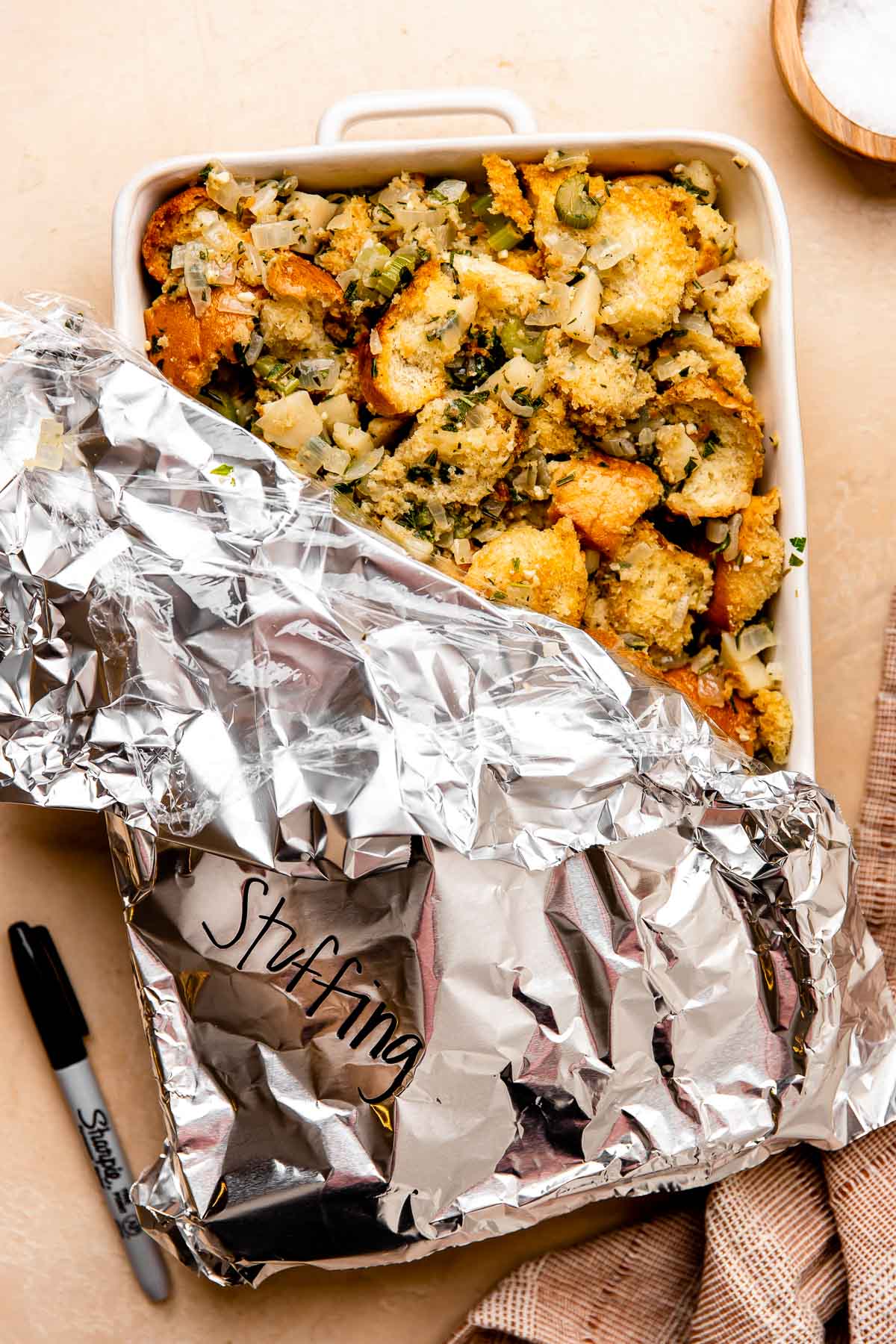 Partially assembled make ahead thanksgiving stuffing fills a large white ceramic baking dish that sits atop a light peach colored textured surface. The baking dish is partially covered with plastic wrap and aluminum foil and the words stuffing have been written on the outside of the foil in black permanent marker. The baking dish is surrounded by a black Sharpie marker, a small wooden pinch bowl filled with kosher salt, and a red muted linen napkin.