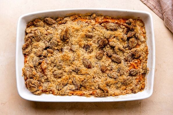 A final make ahead sweet potato casserole for Thanksgiving sits atop a light peach colored textured surface with streusel and maple pecan topping. A muted red linen napkin rests alongside the baking dish.