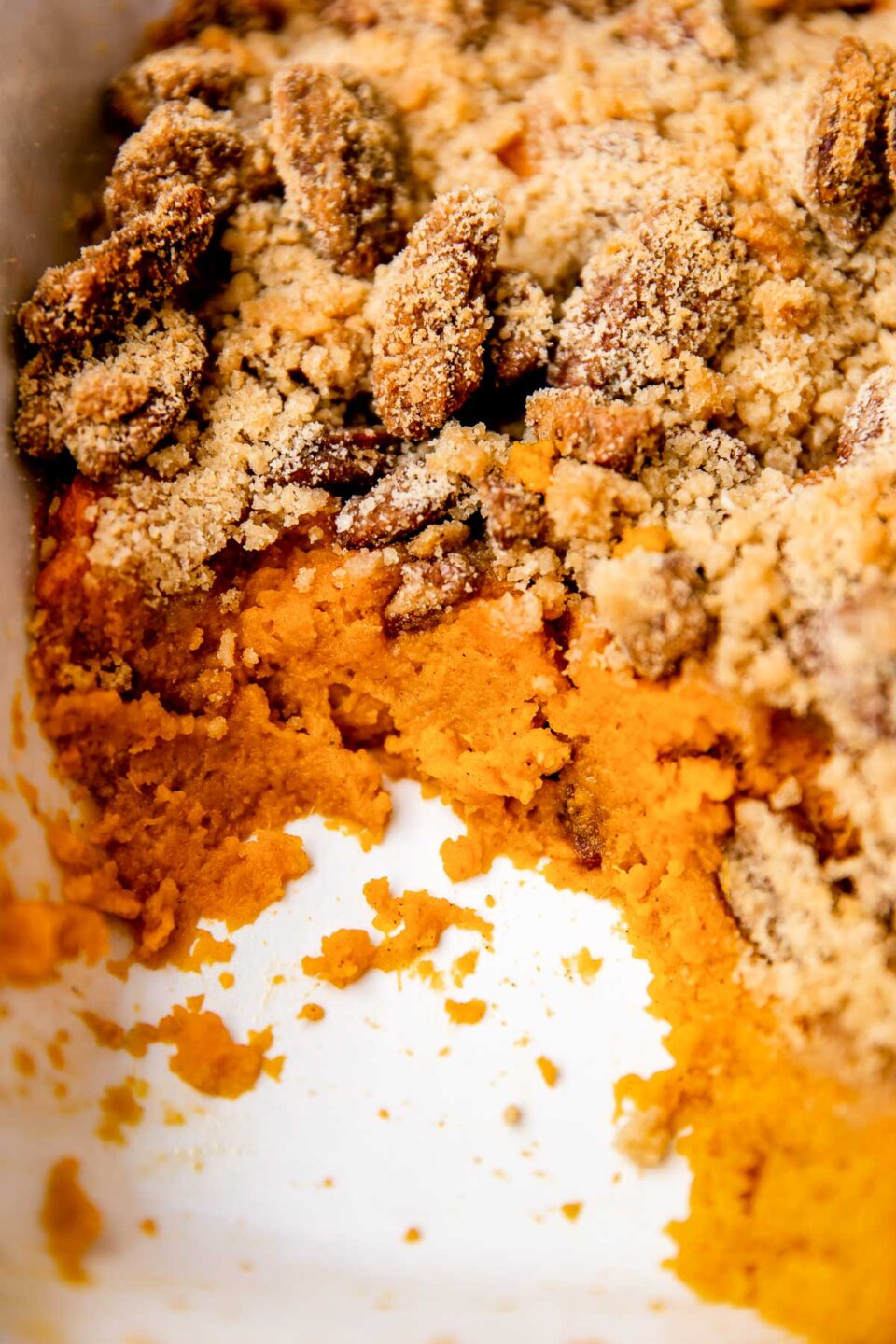 A close up and macro shot of the inside of make ahead thanksgiving sweet potato casserole inside of a large white baking dish. A portion of the make ahead sweet potato casserole has been dished out revealing the layers of sweet potato casserole filling topped with streusel and maple pecan topping.